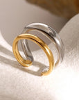 A close-up of a Princess Goes Hollywood zweifarbig Edelstahl Ring on a textured white surface. The verstellbar open ring features intertwined polished gold and silver bands in a minimalist, contemporary design.