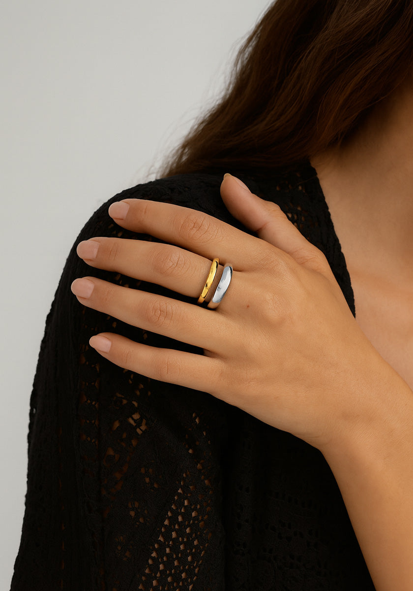 A woman with long brown hair wears a black textured garment by Princess Goes Hollywood. Her hand with neatly manicured nails rests on her chest, displaying a two-tone adjustable stainless steel ring—gold and silver—on her middle finger.