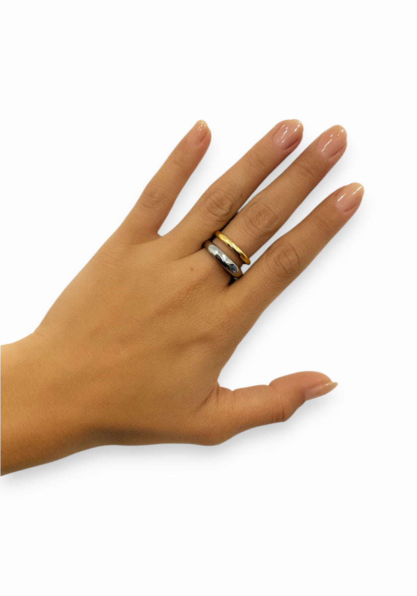 A left hand with light brown skin and manicured nails is shown against a white background, wearing on the ring finger a Princess Goes Hollywood zweifarbig Edelstahl Ring, a gold band, and a silver ring with white stones.