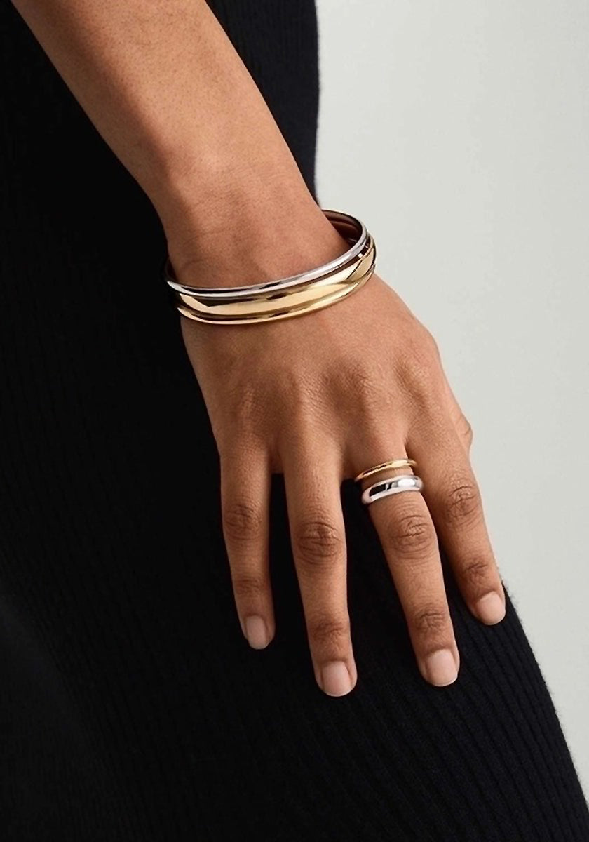A close-up of a hand wearing a zweifarbig Edelstahl Ring by Princess Goes Hollywood and three stacked metallic bangles in gold, silver, and rose gold tones against ribbed black fabric for a minimalist, elegant look.