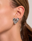 A close-up of a womans ear wearing a large, wavy Edelstahl Ohrstecker with glänzende Oberfläche in organische Form by Princess Goes Hollywood. Her brown hair is tied back, and the neutral background highlights the sculpted earring.