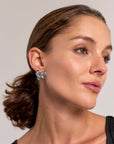 A woman with light skin and brown hair pulled back wears a sleeveless black top and large, shiny stainless steel earrings by Princess Goes Hollywood. She looks slightly to the side with a neutral expression against a plain light background.