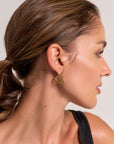A woman with light brown hair in a low ponytail, seen in profile, wears Princess Goes Hollywood Creolen Goldoptik earrings and a black sleeveless top. Her glowing skin stands out against a plain light background.