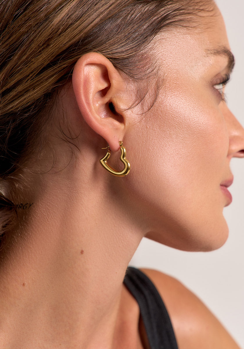 Close-up of a womans right profile showing her ear adorned with Princess Goes Hollywood Creolen in Goldoptik, featuring a modernes Design. She has tan skin, brown hair pulled back, and wears a black top against a softly blurred background.