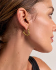 Close-up of a womans right profile showing her ear adorned with Princess Goes Hollywood Creolen in Goldoptik, featuring a modernes Design. She has tan skin, brown hair pulled back, and wears a black top against a softly blurred background.