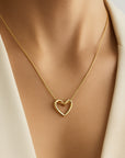 A close-up shows a person with light brown skin wearing a goldene Halskette mit Herzanhänger by Princess Goes Hollywood on a fine chain, styled with a cream or white blazer for an elegant, minimal look.