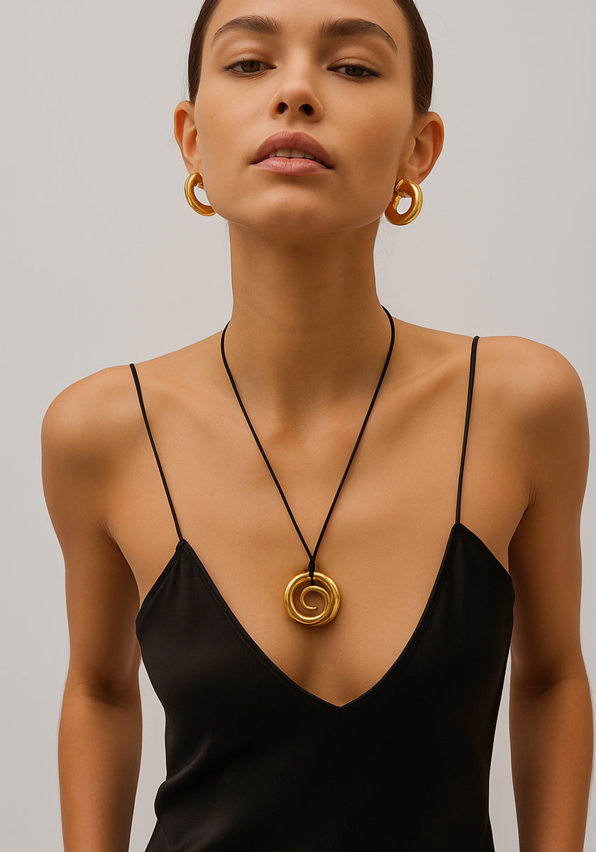A woman in a black spaghetti-strap dress wears large gold hoop earrings and a Princess Goes Hollywood necklace featuring an 18K gold-plated stainless steel pendant. She has straight brown hair pulled back and poses against a light gray background.