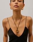 A woman in a black spaghetti-strap dress wears large gold hoop earrings and a Princess Goes Hollywood necklace featuring an 18K gold-plated stainless steel pendant. She has straight brown hair pulled back and poses against a light gray background.