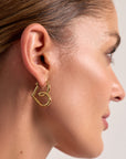 Close-up of a womans right ear wearing Princess Goes Hollywood gold heart earrings with 18K gold plating. Her light brown hair is pulled back, highlighting the modern earring against her smooth skin and a soft, neutral background.