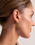 A woman with slicked-back brown hair and glowing skin wears Princess Goes Hollywood gold Herzohrringe with 18K Goldbeschichtung. The ENERGY tattoo behind her ear stands out against a plain, light background.