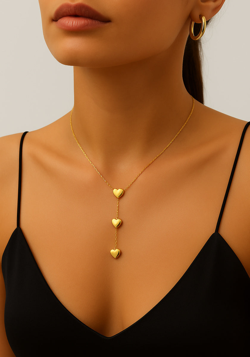 A woman models the Princess Goes Hollywood Zarte Herzkette mit 14K Goldbeschichtung, an elegant stainless steel necklace featuring three descending heart-shaped pendants in a chic vertical design. She pairs it with a black spaghetti strap top and gold hoop earring.