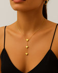 A woman models the Princess Goes Hollywood Zarte Herzkette mit 14K Goldbeschichtung, an elegant stainless steel necklace featuring three descending heart-shaped pendants in a chic vertical design. She pairs it with a black spaghetti strap top and gold hoop earring.