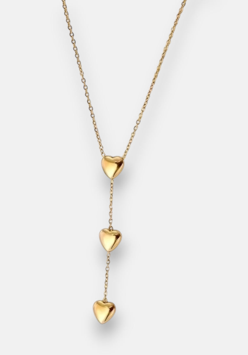 Die goldene Halskette von Princess Goes Hollywood besteht aus einer feinen Edelstahl-Kette mit drei glänzenden Herzanhängern, die vertikal auf einer Seite angeordnet sind und einen eleganten Drop-Effekt erzeugen.