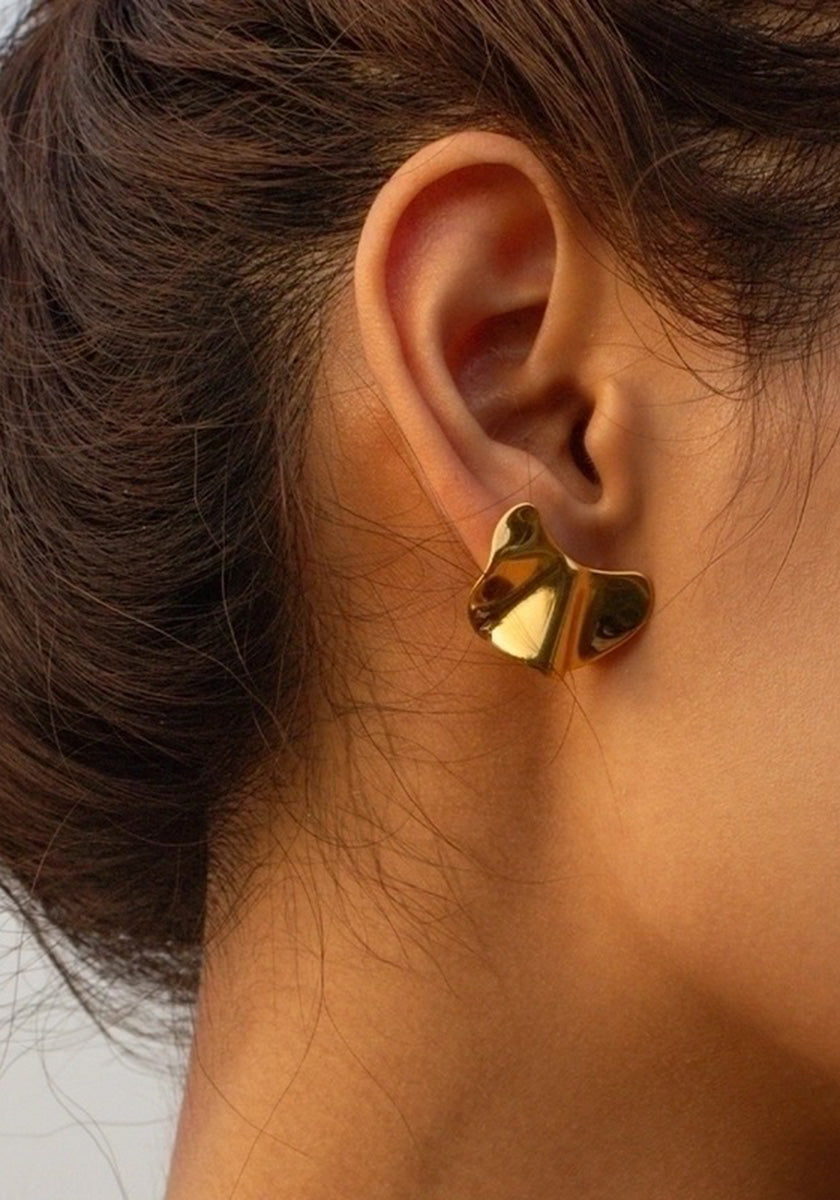 Close-up of a womans ear with dark brown hair tied back, wearing a shiny abstract goldfarben Ohrstecker by Princess Goes Hollywood. The folded-metal earring covers the earlobe, reflects light, and stands out against the neutral background.