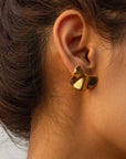 Close-up of a womans ear with dark brown hair tied back, wearing a shiny abstract goldfarben Ohrstecker by Princess Goes Hollywood. The folded-metal earring covers the earlobe, reflects light, and stands out against the neutral background.