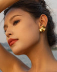 A woman with dark hair pulled back poses outdoors by water, tilting her head and raising one arm. She wears bold gold earrings from Princess Goes Hollywood, with smooth skin, defined brows, long lashes, and red lipstick.