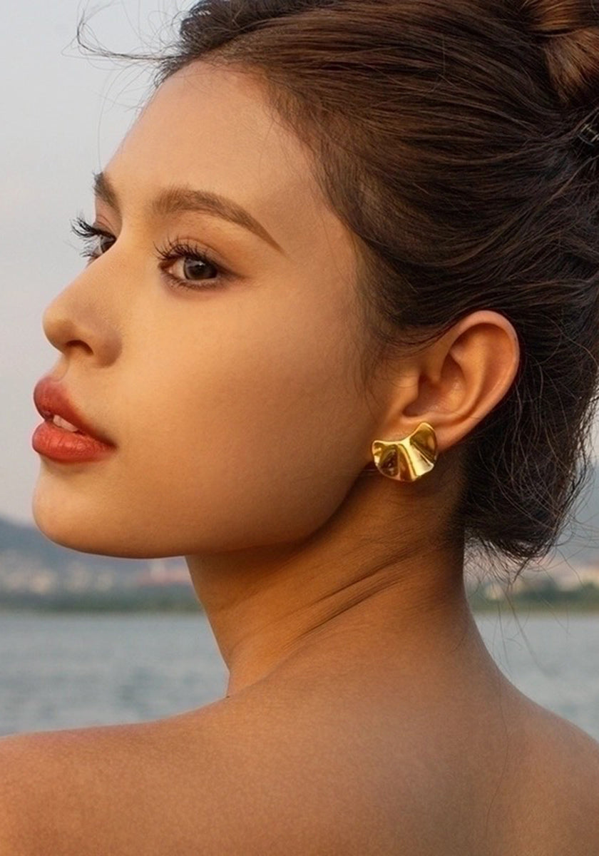 By the water, a woman with smooth skin and light makeup gazes over her shoulder, wearing Princess Goes Hollywood goldfarben Ohrstecker aus Edelstahl shaped like flowers. Her dark hair is up, set against a tranquil outdoor background.