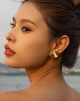 By the water, a woman with smooth skin and light makeup gazes over her shoulder, wearing Princess Goes Hollywood goldfarben Ohrstecker aus Edelstahl shaped like flowers. Her dark hair is up, set against a tranquil outdoor background.