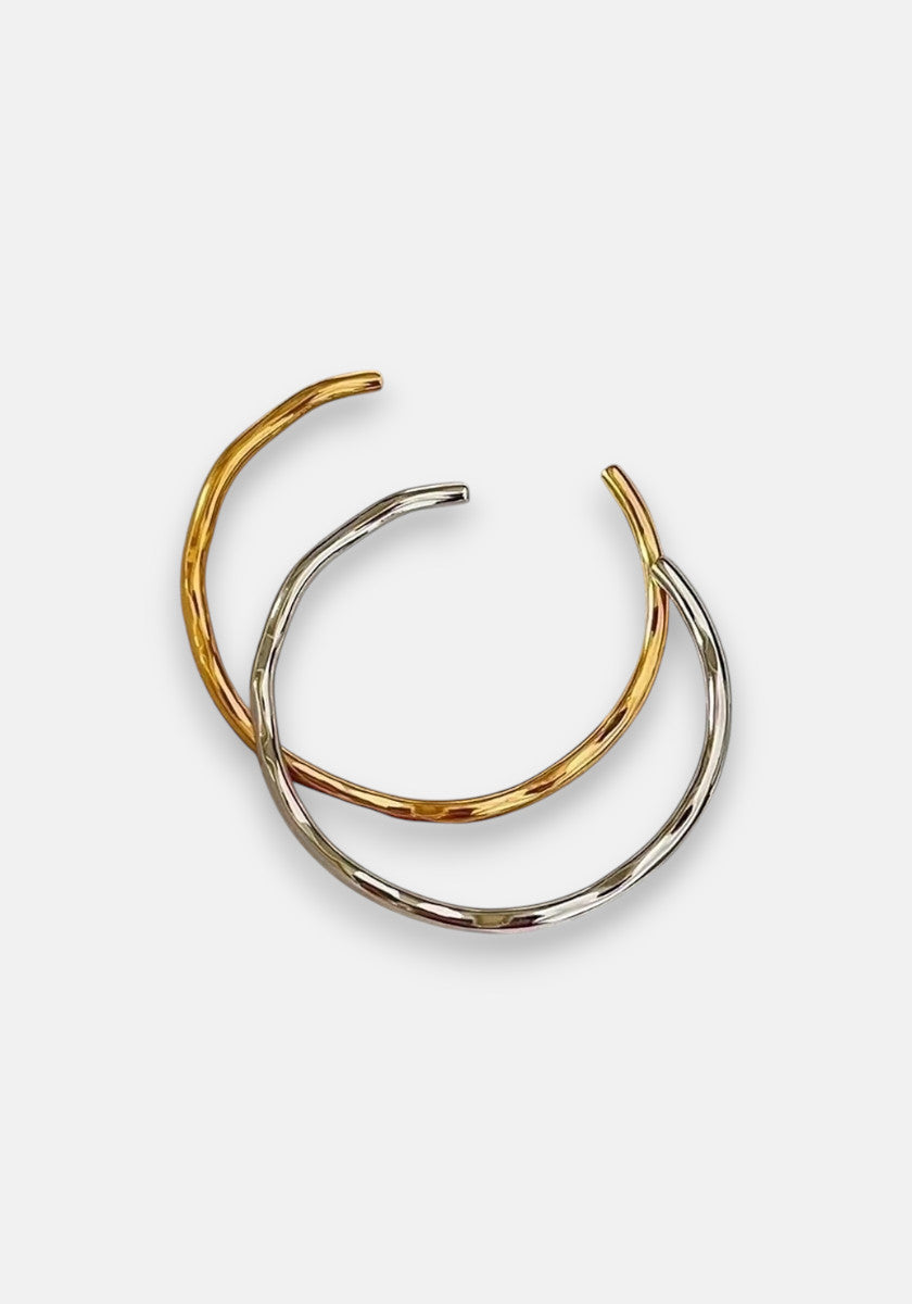 Two thin, open-ended Hoop-Armreifen aus Edelstahl by Princess Goes Hollywood are displayed on white. One has 18K Goldbeschichtung, the other is silver. They partially overlap, creating an intertwined minimalist and elegant look.