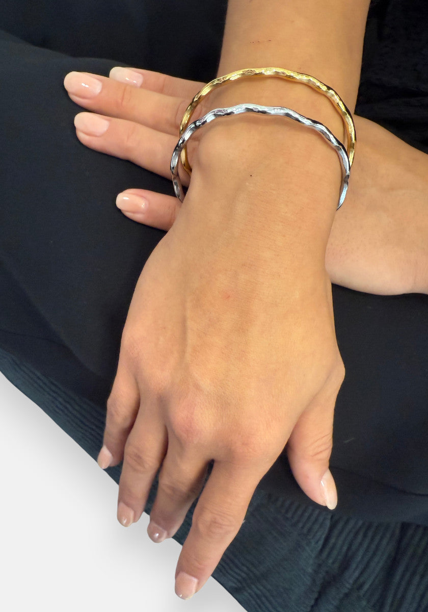 A close-up of hands with manicured nails displaying two thin, wavy Princess Goes Hollywood Armreifen aus Edelstahl mit 18K Goldbeschichtung—one gold, one silver—against a dark fabric background.