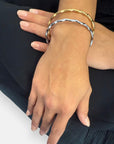 A close-up of hands with manicured nails displaying two thin, wavy Princess Goes Hollywood Armreifen aus Edelstahl mit 18K Goldbeschichtung—one gold, one silver—against a dark fabric background.