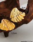 A pair of gold, shell-shaped Ohrstecker by Princess Goes Hollywood rests on textured brown wood. Made from Edelstahl with 18K Goldbeschichtung, one earring measures 3 cm wide, 2.5 cm tall; weight: 12.3g shown in the corner.