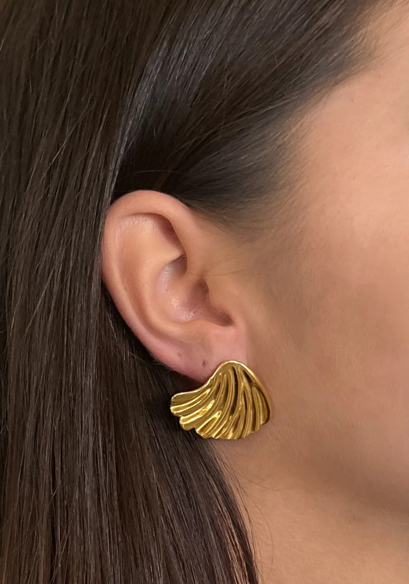 Close-up of a womans ear wearing a bold, shiny Princess Goes Hollywood Ohrstecker crafted from Edelstahl with 18K Goldbeschichtung. The fan-shaped earring features wavy ridges, and her brown hair is tucked to reveal smooth skin.