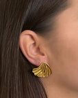 Close-up of a womans ear wearing a bold, shiny Princess Goes Hollywood Ohrstecker crafted from Edelstahl with 18K Goldbeschichtung. The fan-shaped earring features wavy ridges, and her brown hair is tucked to reveal smooth skin.