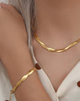 A close-up of a person with light skin and long silver-blonde hair wearing a gold twisted chain necklace and matching Princess Goes Hollywood Gliederarmkette aus Edelstahl mit 18K Goldbeschichtung, plus a cream top with brown polka dots.