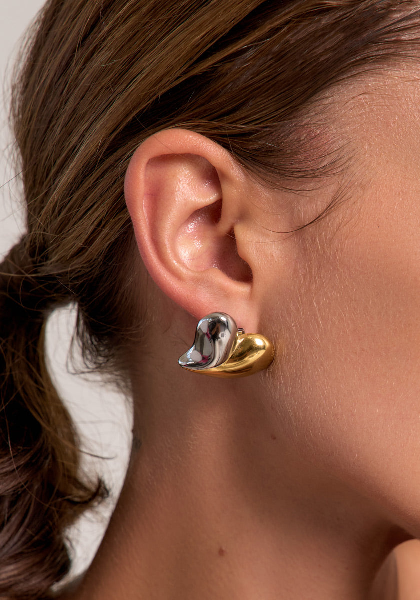 Close-up of a womans ear wearing Princess Goes Hollywoods bold, abstract earring in shiny 18K gold and silver Edelstahl with a wavy bicolor design, contrasting against her smooth skin and pulled-back brown hair.