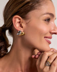 A brown-haired woman in a low ponytail smiles, showing a Princess Goes Hollywood Bicolor-Design abstract earring. She rests her chin on her hands, wearing a matching 18K gold and silver ring. Her skin glows against the light background.