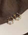 A pair of Princess Goes Hollywood gold stud earrings with three dangling Süsswasserperlen rests on soft brown fabric and a cream surface, highlighting the jewelrys elegance against the textured background.