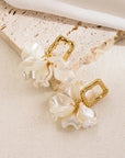 A pair of Princess Goes Hollywood gold Ohrstecker with textured rectangular Edelstahl studs and dangling wavy, iridescent white Kunstperlen, resembling flower petals, displayed on light beige stone with soft white fabric above.