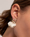 A close-up of a person’s ear adorned with a Princess Goes Hollywood gold Ohrstecker earring featuring a square top, clear gemstone, and white Kunstperlen flower accents. Brown hair is tucked behind the ear, highlighting the elegant piece.