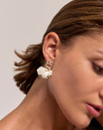 A close-up of a woman’s glowing face in profile, her short brown hair tucked behind her ear, wearing Princess Goes Hollywood Edelstahl Ohrstecker with a chunky gold square and white petal-shaped Kunstperlen.