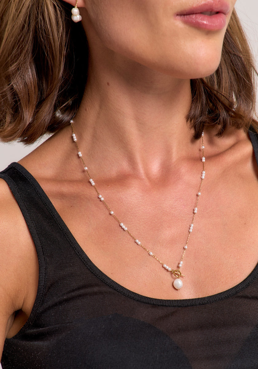 A woman with short brown hair wears a black sleeveless top and a Princess Goes Hollywood Edelstahl Halskette mit Süsswasserperlen: delicate gold necklace with evenly spaced pearls, a central large pearl pendant, and matching pearl earring.