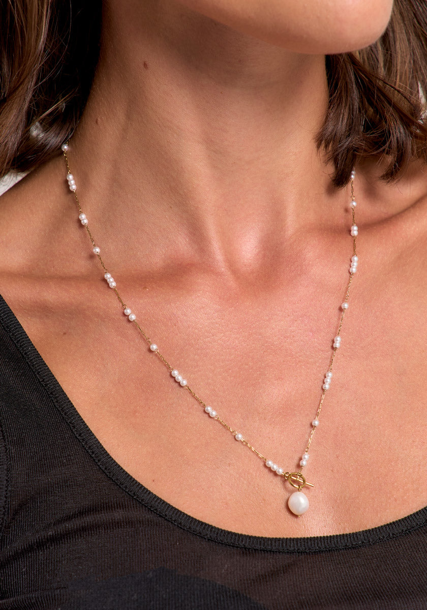 A close-up of a woman wearing a Princess Goes Hollywood Edelstahl Halskette with Süsswasserperlen, gold clasp, and round pearl pendant. She has short brown hair and wears a black sheer top.