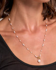 A close-up of a woman wearing a Princess Goes Hollywood Edelstahl Halskette with Süsswasserperlen, gold clasp, and round pearl pendant. She has short brown hair and wears a black sheer top.