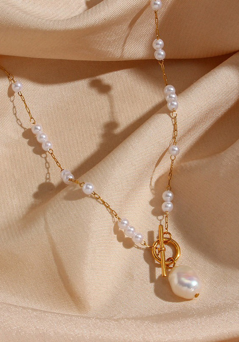 A Princess Goes Hollywood gold chain necklace with Süsswasserperlen rests on beige satin. The Edelstahl Halskette has 18K Goldbeschichtung, a toggle clasp, and a large pearl pendant, with soft shadows adding an elegant touch.