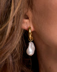 A close-up of a womans ear wearing a Princess Goes Hollywood Creolen Edelstahl hoop earring with a large, irregular Süsswasserperle pendant. Her brown hair frames the 18K goldbeschichtete earring, highlighting its elegant design.