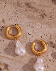 A pair of Princess Goes Hollywood Creolen Edelstahl hoop earrings with 18K Goldbeschichtung, each featuring an irregular baroque-style Süsswasserperle, displayed on a textured beige stone with natural holes and grooves.