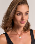 A woman with shoulder-length brown hair wears a black tank top and a Princess Goes Hollywood gold-plated necklace with a round white baroque pearl pendant. She has natural makeup, brown eyes, a slight smile, and stands against a plain light background.
