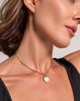 A close-up of a woman’s neck and jawline reveals her wearing a Princess Goes Hollywood gold-plated necklace with a large baroque pearl pendant. She has glowing skin, a black sleeveless top, and brown hair swept to the side.