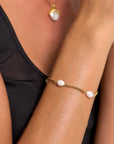 A close-up of a person with light brown skin wearing a black sleeveless top, highlighting Princess Goes Hollywood’s gold-plated bracelet with freshwater pearls and a matching gold necklace featuring a large white pearl.