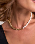 A close-up of a woman’s neck wearing a Princess Goes Hollywood necklace: half 18K Goldbeschichtung gedrehte Gliederkette, half weiße Süsswasserperlen. She has shoulder-length brown hair, a black asymmetrical top, and a neutral background.