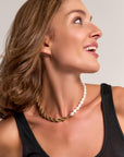 A smiling woman with wavy brown hair looks right, wearing a sleeveless black top and a Princess Goes Hollywood Halskette that combines Süßwasserperlen with 18K-Goldbeschichtung. The light background highlights her joyful, confident expression.
