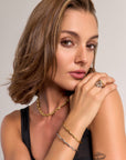A woman with chin-length brown hair poses in a black sleeveless top, highlighting Princess Goes Hollywood jewelry: a statement necklace, gold and silver shell-link bracelets with 18K gold plating, and a statement ring. A small forearm tattoo is visible.
