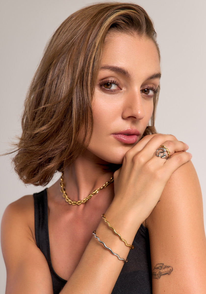 A woman with short brown hair models a Princess Goes Hollywood minimalist jewelry set: a chain necklace, stainless steel bangles, and an 18K gold-plated ring. She wears natural makeup and confidently displays her paper airplane tattoo.