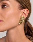 Close-up of a woman’s face and ear, highlighting a large, gold earring from Princess Goes Hollywood. Its abstract design, shiny surface, and unique folded form are accentuated by her smooth skin and low ponytail.