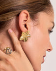 Close-up of a womans ear wearing large, gold floral stud earrings by Princess Goes Hollywood. Her brown hair is tucked back, and she gently touches her earlobe while wearing a stainless steel heart-shaped ring. The background is neutral.